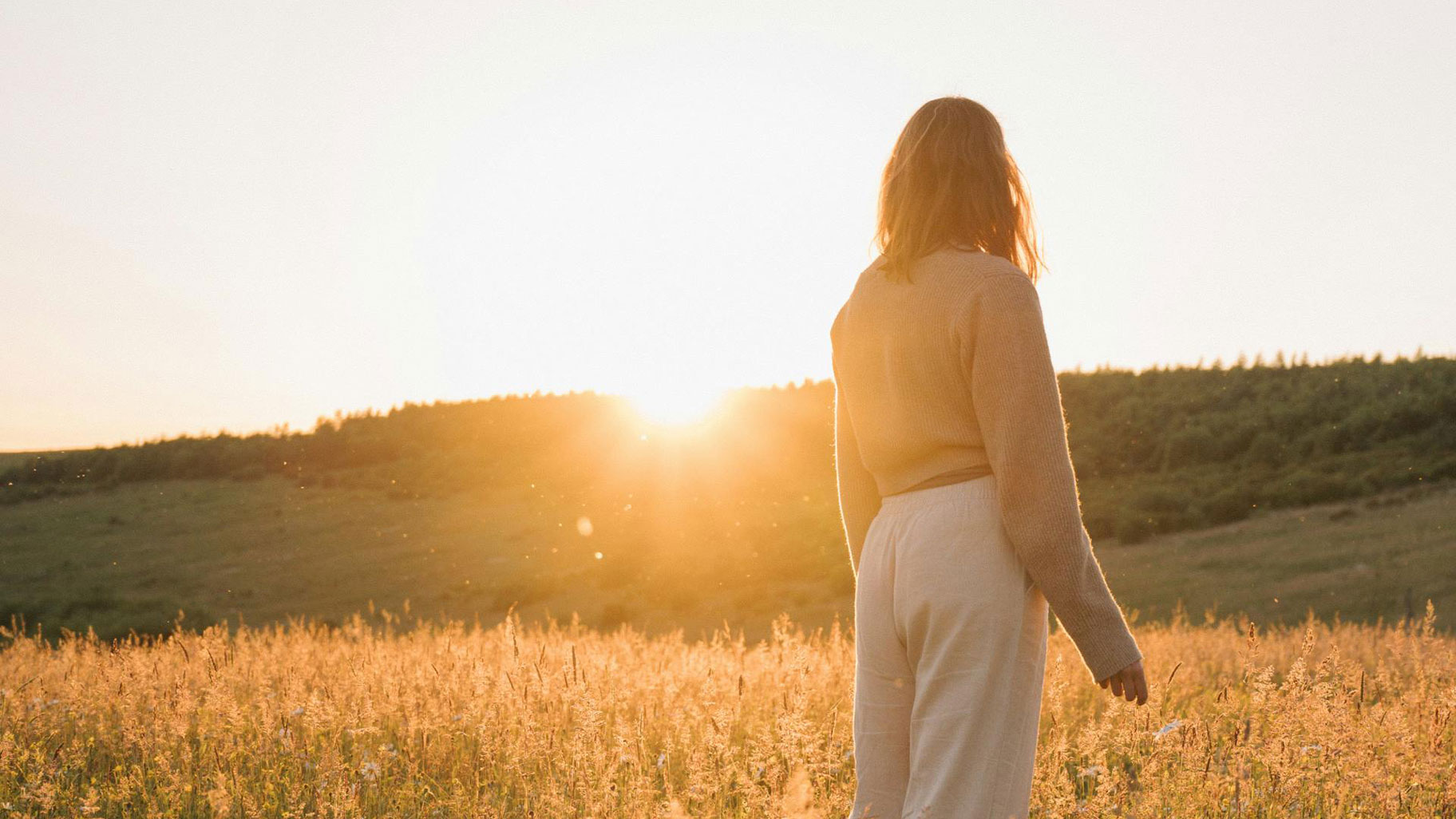 Landscape photo of a woman standing in a field, facing the sun, with a scenic natural background.